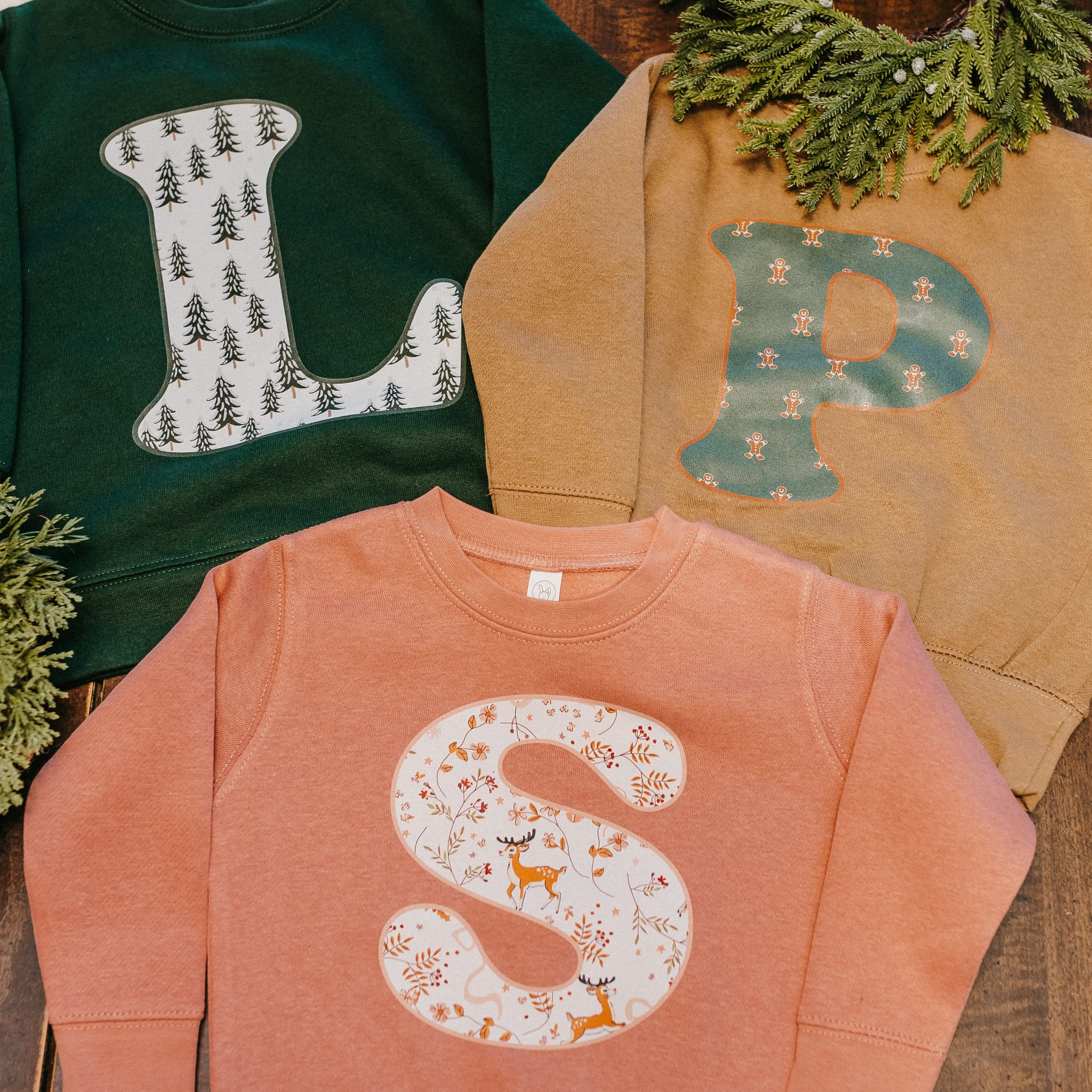 Child sweaters in forest, coyote brown, and mauvelous with Christmas patterned initials on front, shown on flat lay – Little Mama Shirt Shop