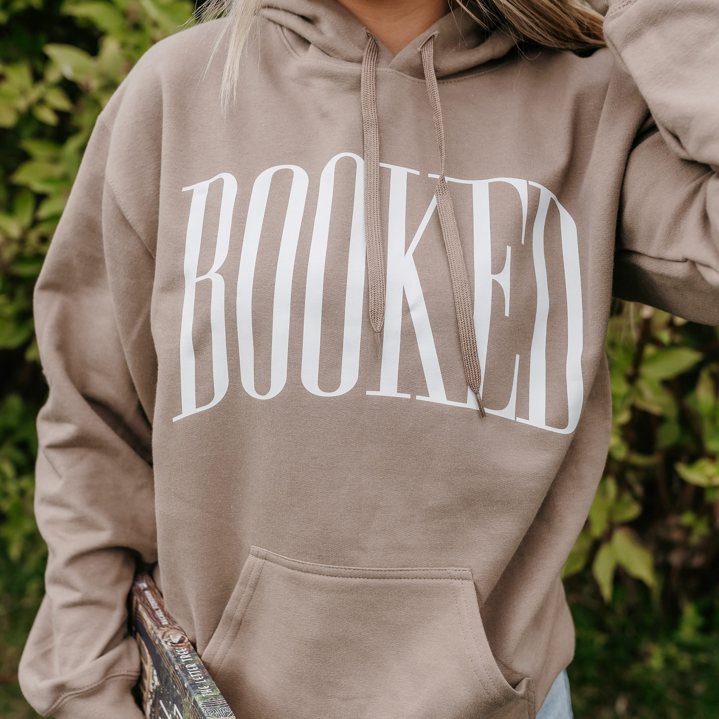 Unisex hoodie in paragon with 'BOOKED' arched text close up on model – Little Mama Shirt Shop